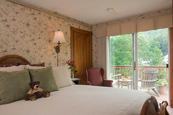 Guest room with king sleigh bed with ivory bedding, burgundy wing chair in front of door, sliding glass doors with view of balcony with table & chairs