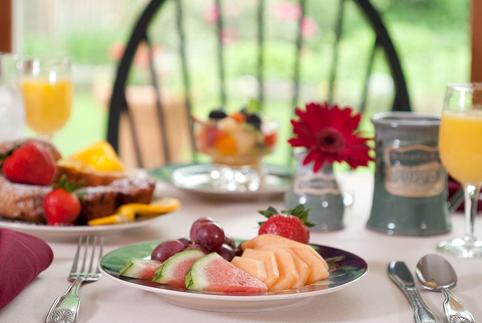 plate of fruit with red flower in vase, coffee mug, fruit cup, and black Windsor chair in background