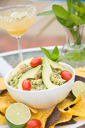 White bowl of guacamole with cherry tomatoes, chips, lime in foreground, drink in background