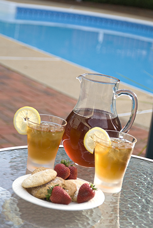 Iced tea pitcher and glasses with plate of cookies and strawberries on glass table, pool in background