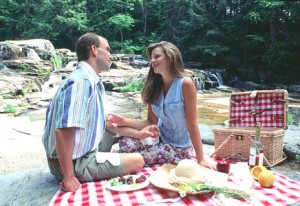 couple sitting on red & white plaid cloth beside waterfall behind them with picnic food in front, picnic basket beside them