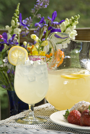 Goblet & pitcher of lemonade with wildflower bouquet behind & strawberries on plate