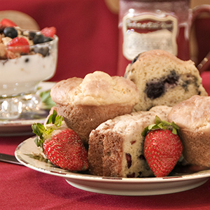 plate of muffins garnished with strawberries with bowl of yoghurt & berries & coffee mug in backgroun