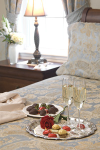 Chocolate strawberries, champagne, chocolates on bed with table & lamp in background