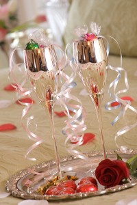 Silver champagne flutes with chocolates & ribbons on silver tray on rose petal strewn bed