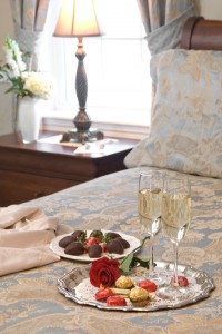 Bed with blue bedding with silver tray with chocolates, red rose, champagne in glasses, plate of chocolates behind.