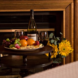 round table with plate of fruit, red wine glasses & bottle, sunflowers to right, fireplace behind table